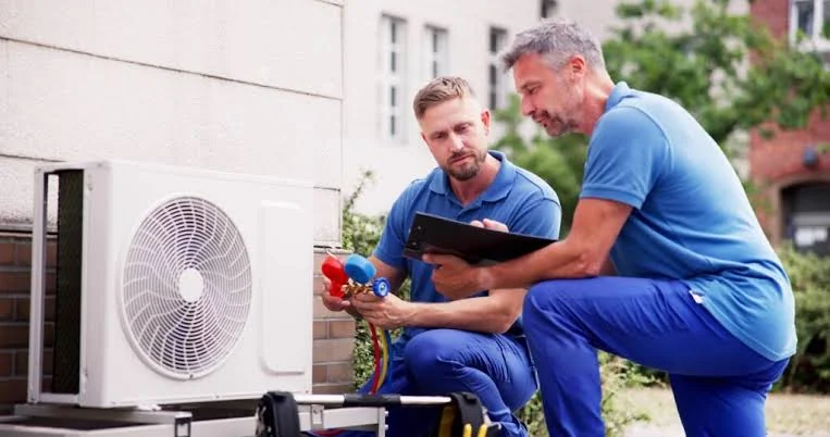 Two HVAC technicians checking an outdoor unit with gauges and a tablet